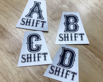 May include: Four white triangular stickers with black lettering. Each sticker has a letter, A, B, C, or D, and the word "SHIFT" underneath.