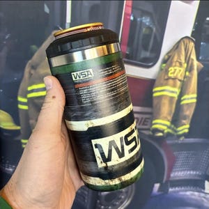 May include: A cylindrical stainless steel beverage cooler with a black plastic rim and a green, black, and white wrap featuring the text WSA. The design includes a weathered, distressed texture and printed warning labels on the upper section.