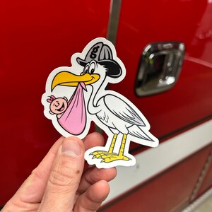 Puede incluir: Una cig&uuml;e&ntilde;a blanca con un sombrero de bombero negro y gris, sosteniendo una manta de beb&eacute; rosa con un beb&eacute; peque&ntilde;o dentro. La cig&uuml;e&ntilde;a sostiene al beb&eacute; en su pico. La pegatina est&aacute; en una superficie roja.