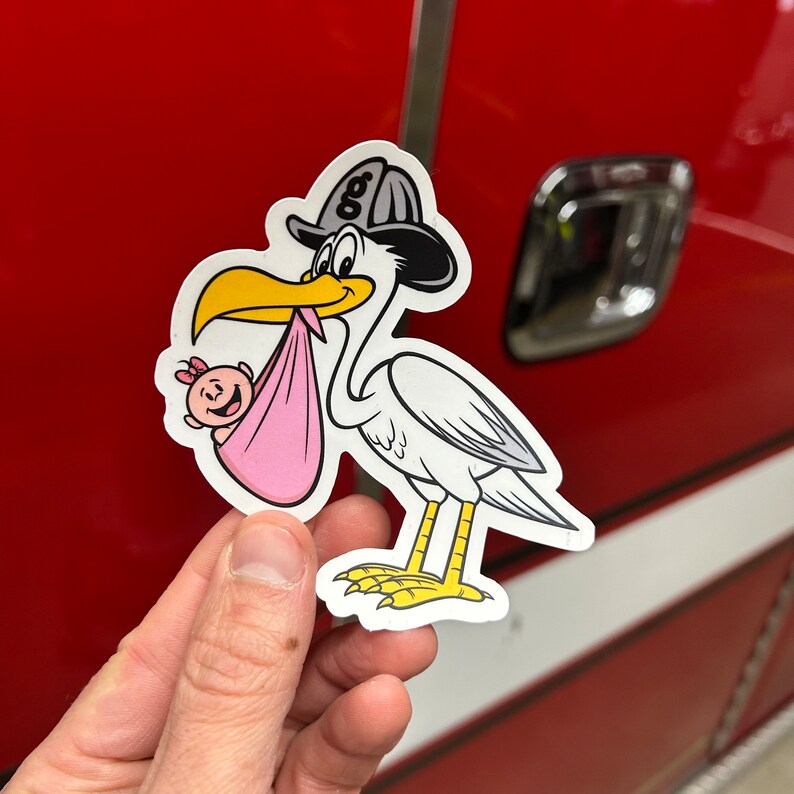 Puede incluir: Una cig&uuml;e&ntilde;a blanca con un sombrero de bombero negro y gris, sosteniendo una manta de beb&eacute; rosa con un beb&eacute; peque&ntilde;o dentro. La cig&uuml;e&ntilde;a sostiene al beb&eacute; en su pico. La pegatina est&aacute; en una superficie roja.