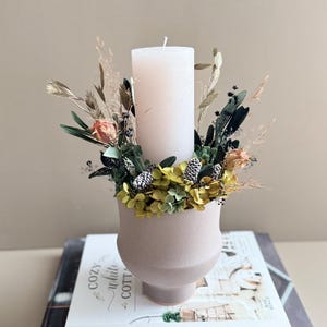 Op de afbeelding: Een witte pilaar kaars staat in een zachtroze vaas gevuld met gedroogde bloemen en groen. De vaas staat op een stapel boeken, waaronder een met de titel "Country and Cozy".