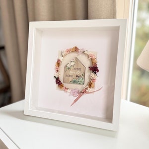 May include: A white frame containing a pink and white floral wreath surrounding a wooden house with the text "Home Sweet Home".