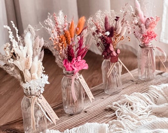 May include: Four small glass jars filled with dried flowers and greenery. Each jar has a small tag that says "Merry Christmas!" The jars are arranged on a wooden surface with a white knitted blanket in the foreground.