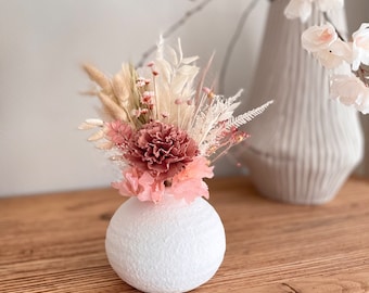 May include: A white ceramic vase with a textured surface holds a bouquet of dried flowers. The bouquet includes pink carnations, beige pampas grass, and white dried flowers.