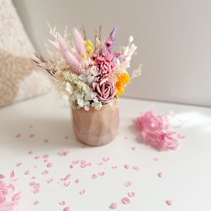 May include: A small, peach-colored vase holds a vibrant arrangement of dried flowers. The bouquet features pink, purple, yellow, and white blooms, with a prominent rose. Scattered pink petals add a delicate touch. A cozy, neutral-toned background complements the floral display.