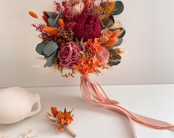 May include: A dried flower bouquet in shades of orange, pink, and red with a long pink ribbon. The bouquet includes dried roses, eucalyptus, and other dried flowers. A matching buttonhole is also included.