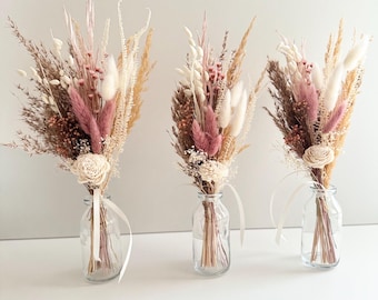 May include: Three small glass vases filled with dried flower arrangements. Each vase contains a mix of dried grasses, pampas grass, and pink and white flowers.