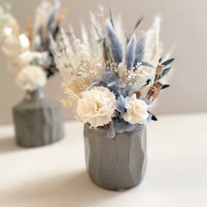 Op de afbeelding: Een grijze keramische vaas met een getextureerd oppervlak bevat een boeket gedroogde bloemen. Het boeket bevat witte anjers, blauwe gedroogde bloemen en wit pampasgras.