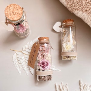 Op de afbeelding: Drie glazen flessen met kurken, elk gevuld met gedroogde bloemen en decoratieve elementen. Eén fles heeft een roze roos, terwijl andere witte en roze bloemstukken bevatten. Elke fles heeft een klein label.