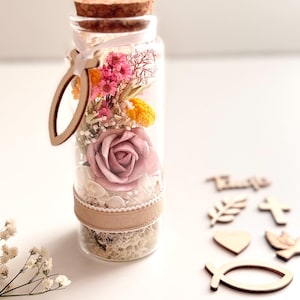 Op de afbeelding: Een heldere glazen fles gevuld met gedroogde bloemen, waaronder een roze roos, en decoratieve elementen. De fles is afgesloten met een kurk en een houten bedel. Extra houten uitsparingen van religieuze symbolen liggen in de buurt verspreid.
