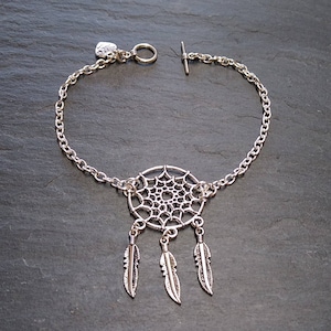 Puede incluir: Pulsera plateada con un colgante de atrapasueños y tres colgantes de plumas. La pulsera tiene un diseño de cadena y un cierre de palanca. El atrapasueños tiene un diseño de telaraña circular.