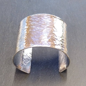 May include: A wide, hammered silver cuff bracelet. The bracelet has a textured surface with a series of small indentations. The open cuff design allows for easy wear. This statement piece is a bold accessory.