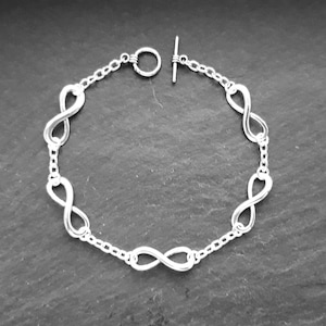 May include: A silver infinity symbol bracelet with a toggle clasp. The bracelet has five infinity symbols connected by a chain.