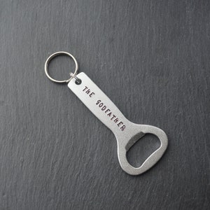 May include: A silver bottle opener with a key ring. The words "THE GODFATHER" are stamped on the handle. The opener is designed with a curved shape for easy use.