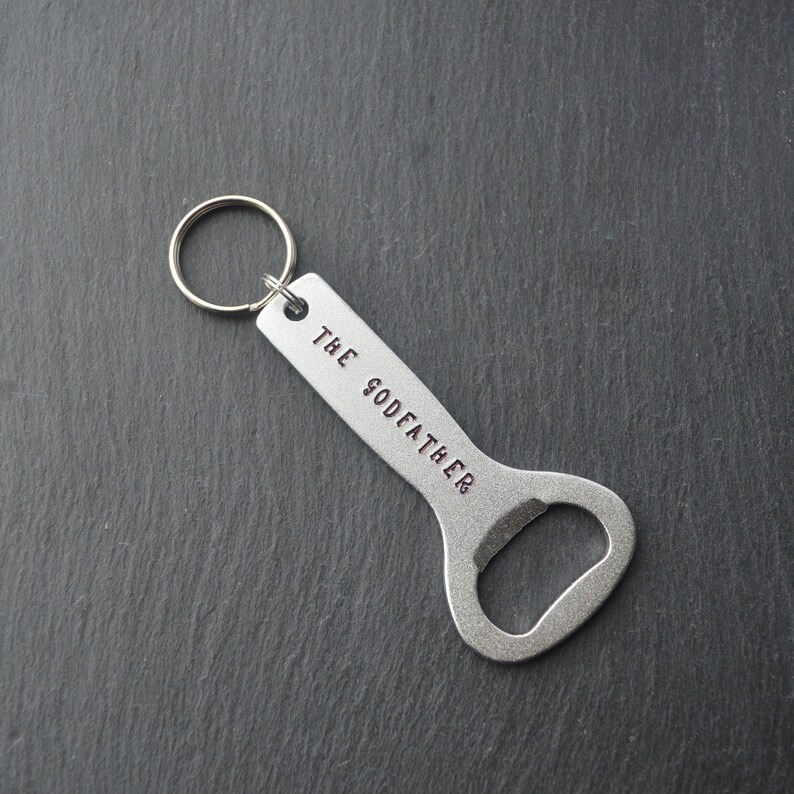 May include: A silver bottle opener with a key ring. The words "THE GODFATHER" are stamped on the handle. The opener is designed with a curved shape for easy use.