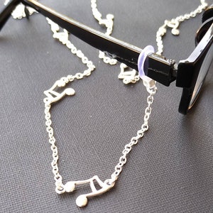 Puede incluir: Cadena de plata con dijes de notas musicales unidos a un par de gafas negras. La cadena está asegurada a las gafas con un anillo de plástico transparente.
