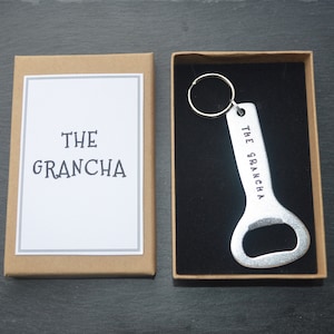 May include: A silver bottle opener with a keychain ring. The bottle opener has the text "THE GRANCHA" engraved on it. The bottle opener is in a brown box with a white label that says "THE GRANCHA".