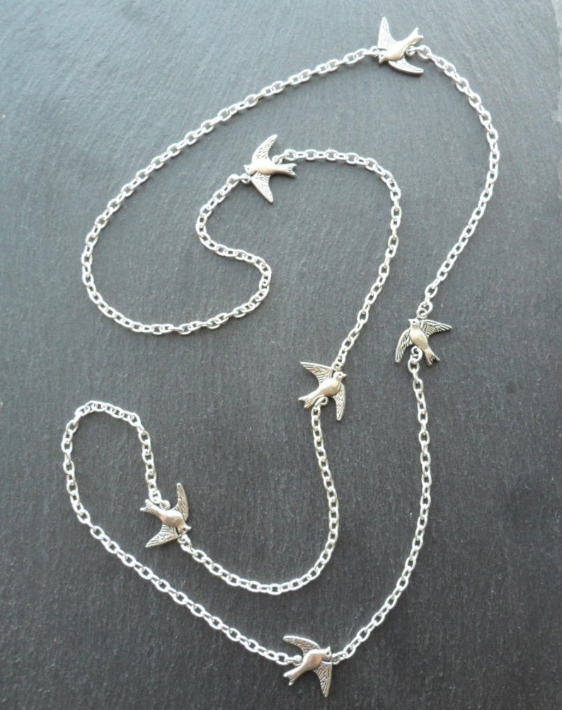May include: A silver chain necklace with five silver bird charms. The chain is long and has a delicate design. The birds are facing different directions and are in flight.