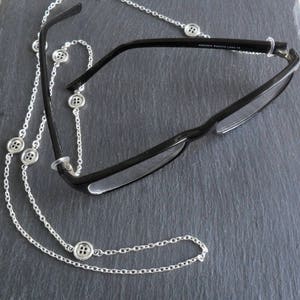 May include: A silver chain with small buttons attached, used to hold glasses. The chain is looped around the arms of a pair of black eyeglasses.