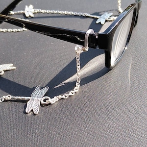 May include: A silver chain with dragonfly charms attached to black eyeglasses. The chain is designed to hold the glasses around the neck.