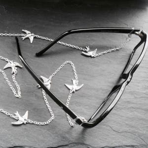 May include: A silver chain with small bird charms attached to black eyeglasses. The chain is designed to hold the eyeglasses around the neck.