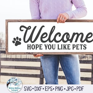 Peut inclure: Une pancarte en bois avec le texte "Welcome. Hope you like pets." en lettres noires. La pancarte est tenue par une personne portant une chemise violette et un jean bleu. Deux empreintes de pattes se trouvent de chaque côté du texte.