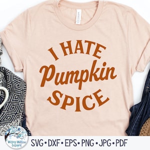 May include: A light pink t-shirt with the text "I HATE Pumpkin Spice" in orange lettering. The shirt is laid flat on a white background with a scarf, a mug, and a pair of jeans.
