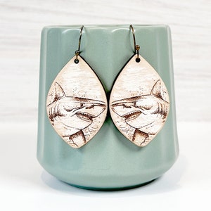 May include: A pair of teardrop-shaped wooden earrings featuring a laser-engraved shark illustration. The earrings hang from dark metal hooks and display a detailed shark head with visible teeth and fins, rendered in a dark brown wood-burned style.