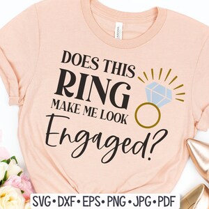 May include: A peach-colored t-shirt with the text "Does this ring make me look engaged?" in black. A gold ring with a blue diamond is pictured next to the text.