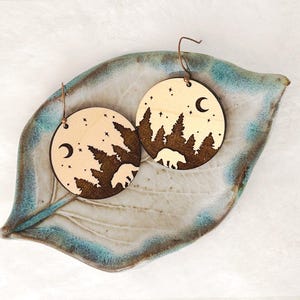May include: Two circular wooden earrings featuring a laser-engraved forest scene with a bear, pine trees, stars, and a crescent moon. The earrings have metal hooks. Text on the right side reads: Bear EARRINGS, Glowforge Tested LASER CUT FILE.