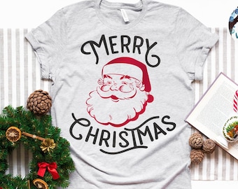 May include: A light gray short-sleeved t-shirt featuring a red and black graphic of Santa Claus with the text Merry Christmas. The shirt is displayed alongside a green pine wreath with red bows and dried orange slices, and an open book with candy canes.