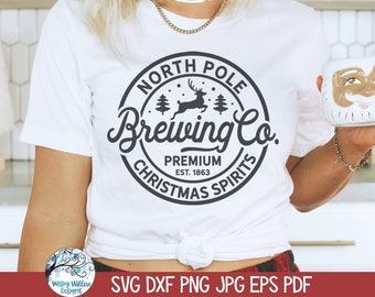 May include: A white t-shirt featuring a black circular graphic with the text North Pole Brewing Co. Premium Est. 1863 Christmas Spirits, accompanied by a reindeer and pine tree illustration. The shirt is tied at the waist and worn with a Santa mug.