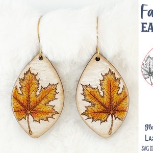 Könnte beinhalten: Ein Paar tropfenförmige Holzohrringe mit einer detaillierten Illustration eines Ahornblatts in Orange- und Gelbtönen. Der Text Fall Leaf EARRINGS, Glowforge Tested LASER CUT FILE und SVG DXF PNG JPG EPS PDF AI ist sichtbar.