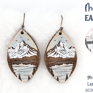 Könnte beinhalten: Tropfenförmige Holzohrringe mit einer lasergravierten Berglandschaft, einem See und Tannenbäumen. Das Design enthält weiße und blaue Farbakzente. Der Text lautet Mountain EARRINGS, Wispy Willow Designs, Glowforge Tested LASER CUT FILE, SVG DXF PNG JPG EPS PDF AI.