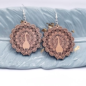 May include: A pair of wooden peacock earrings with a mandala design. The earrings are brown and have silver hooks. The earrings are displayed on a blue feather-shaped dish.