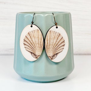 May include: Oval-shaped wooden earrings featuring a detailed laser-engraved seashell design on a light wood surface with dark metal hooks. The text Seashell Earrings, Glowforge Tested Laser Cut File, SVG DXF PNG JPG EPS PDF AI is visible.