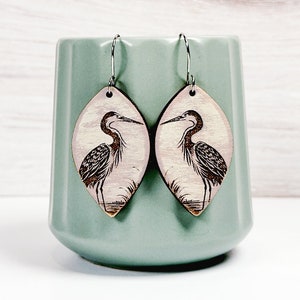 May include: A pair of teardrop-shaped wooden earrings featuring a detailed black ink illustration of a heron standing in water. The earrings have silver-toned metal hooks. The text "HERON EARRINGS" and "Glowforge Tested LASER CUT FILE" is visible.