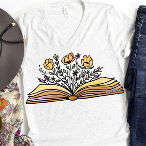 Op de afbeelding: Een wit T-shirt met een ontwerp van een open boek met gele bloemen die eruit groeien. De tekst "Wispy Willow" is op het boek gedrukt.