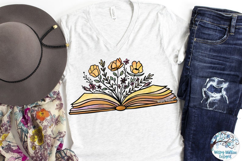 Op de afbeelding: Een wit T-shirt met een ontwerp van een open boek met gele bloemen die eruit groeien. De tekst "Wispy Willow" is op het boek gedrukt.