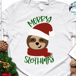 Può includere: Una maglietta grigia con una grafica verde e rossa di un bradipo che indossa un cappello da Babbo Natale e una sciarpa. Il testo "Merry Slothmas" è scritto in verde sotto il bradipo.