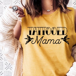 May include: A mustard yellow sleeveless top featuring the black text "TATTOOED Mama" printed on the chest. The design includes two black swallow illustrations flanking the word "Mama." The top is worn with a cream-coloured knit cardigan.