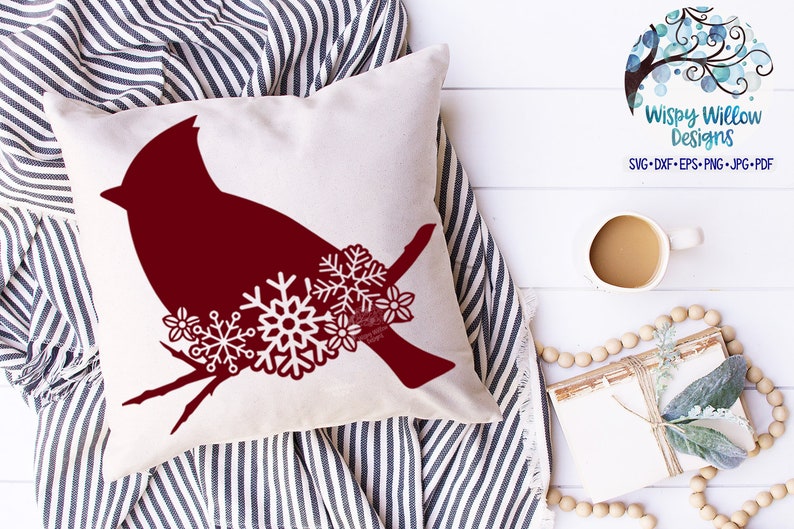 May include: A red cardinal silhouette with snowflakes and berries on a white pillow. The pillow is on a striped fabric background.