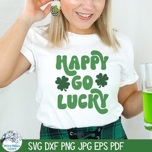 Op de afbeelding: Een wit T-shirt met korte mouwen en de groene tekst "HAPPY GO LUCKY" met twee klaverbladiconen. Het shirt wordt gedragen met een groen geruite rok en een glazen mok gevuld met een felgroene vloeistof, inclusief een bierpul-oorbel.