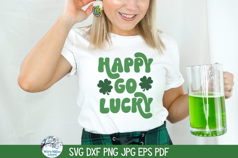 Op de afbeelding: Een wit T-shirt met korte mouwen en de groene tekst "HAPPY GO LUCKY" met twee klaverbladiconen. Het shirt wordt gedragen met een groen geruite rok en een glazen mok gevuld met een felgroene vloeistof, inclusief een bierpul-oorbel.