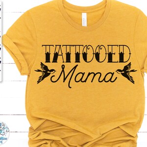 May include: A mustard yellow short-sleeve t-shirt featuring the black text "TATTOOED Mama" centred on the chest. The word "TATTOOED" is in a bold, blocky font, while "Mama" is in a cursive script. Two black swallow illustrations flank the text.