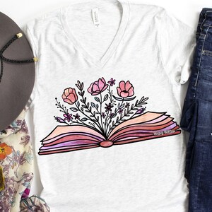 Op de afbeelding: Een wit T-shirt met een roze en wit bloemenontwerp van bloemen die uit een open boek groeien. De tekst "Wispy Willow" is op het boek gedrukt.