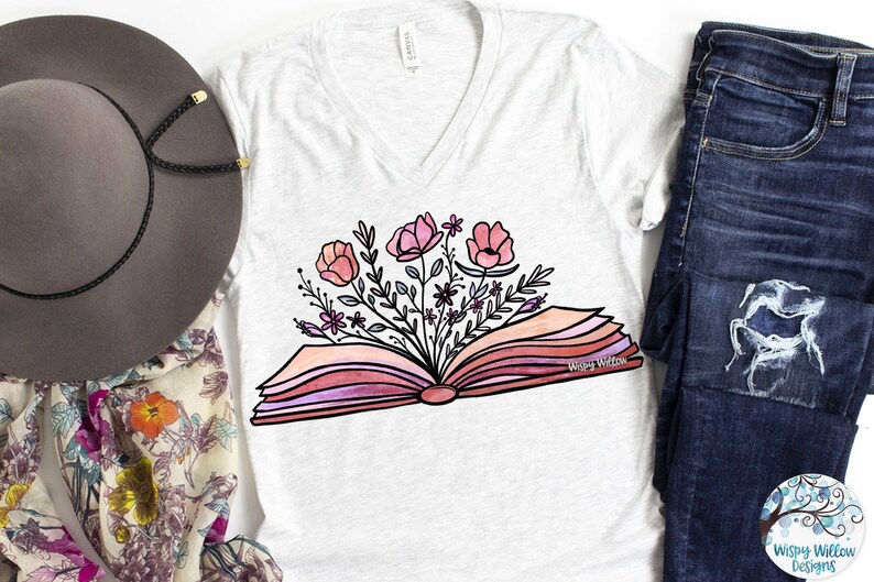 Op de afbeelding: Een wit T-shirt met een roze en wit bloemenontwerp van bloemen die uit een open boek groeien. De tekst "Wispy Willow" is op het boek gedrukt.