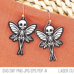 May include: Two skeleton fairy earrings with black and white wings. The earrings are hanging from a brown fabric background.