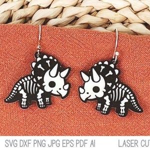 May include: A pair of dangle earrings featuring a black and white triceratops dinosaur skeleton design. Each earring is suspended from a silver-toned metal hook. The dinosaur graphic shows a stylized skeletal structure with a large frill and horns.
