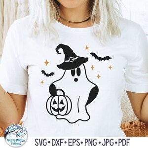 May include: A white short-sleeved t-shirt featuring a black graphic of a ghost wearing a witch hat and holding a jack-o'-lantern. The design includes small orange four-pointed stars and two black bats flying on either side of the ghost figure.
